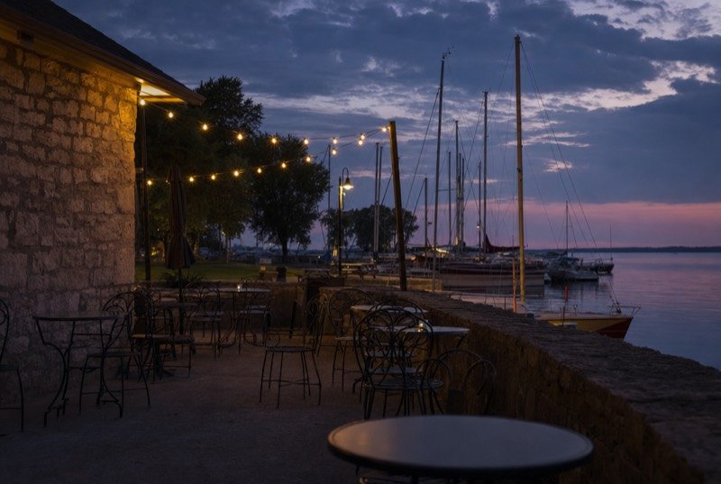 Empty bistro tables on a stone-edged waterfront patio at dusk, with sailboat masts and string lights behind