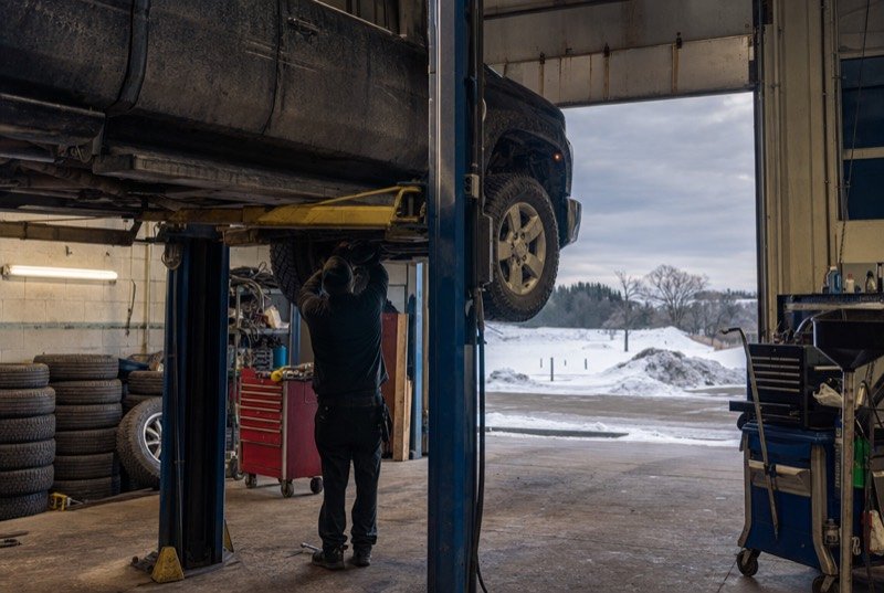 A raised pickup in an independent repair bay, a stack of winter tires against the wall and snow beyond the door