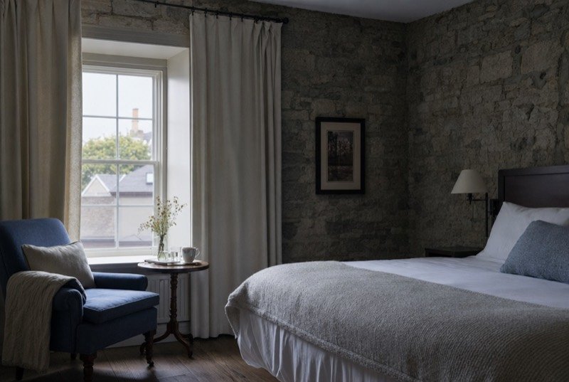 A quiet guest room with an exposed grey stone wall, a made bed and a reading chair in morning light