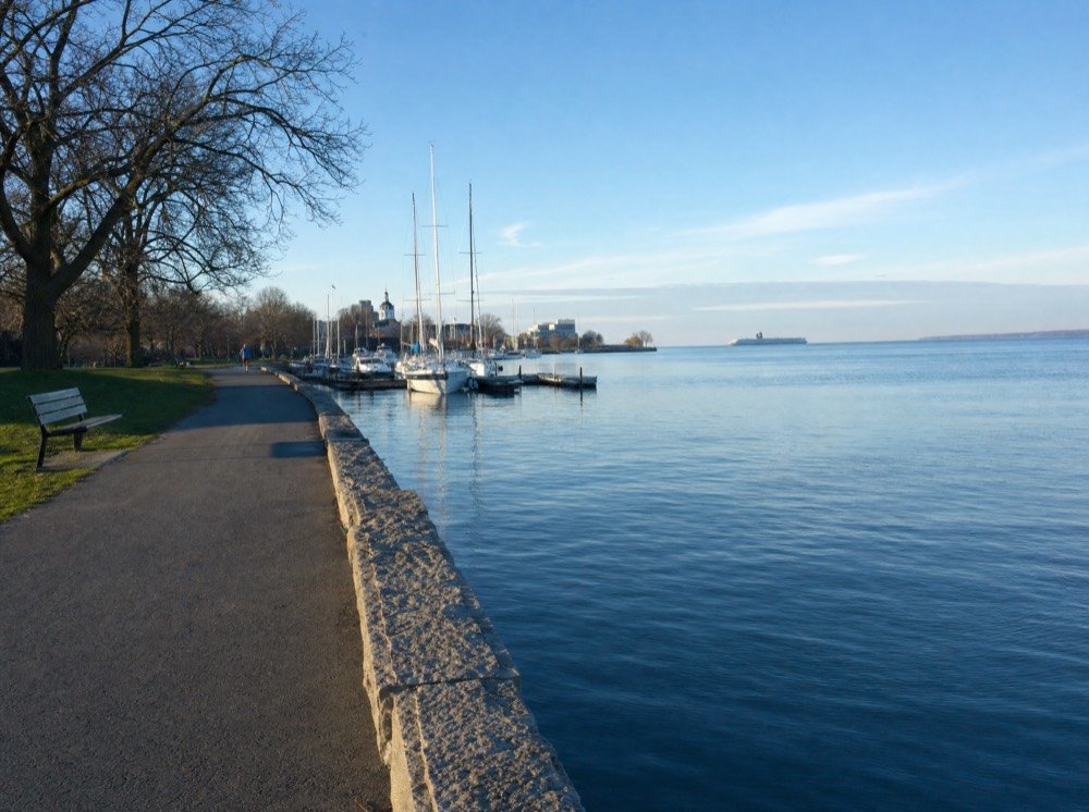 A harbour-side path beside a low stone wall, sailboats at their moorings and a bench in morning sun